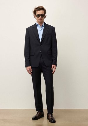 Mann in schwarzem Anzug, hellblauem Hemd, dunkler Sonnenbrille und polierten braunen Lederschuhen steht vor einem einfarbigen hellen Hintergrund.