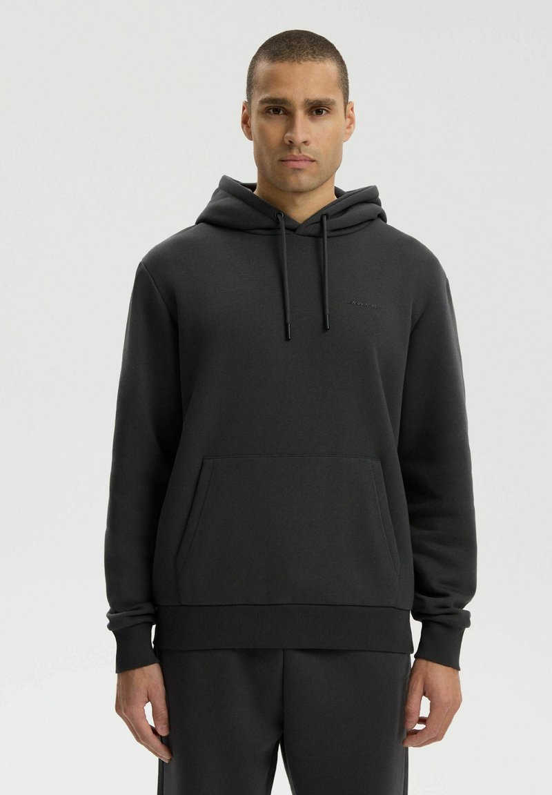 Mężczyzna ubrany w jednolitą czarną bluzę z kapturem i przednią kieszenią, stojący na neutralnym tle, zwrócony przodem z neutralnym wyrazem twarzy.