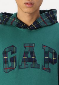 Hanorac verde cu un căptușeală în carouri albastre în glugă. Prezintă un logo brodat mare "GAP" într-un model de carouri asortat. Textură de material neted.