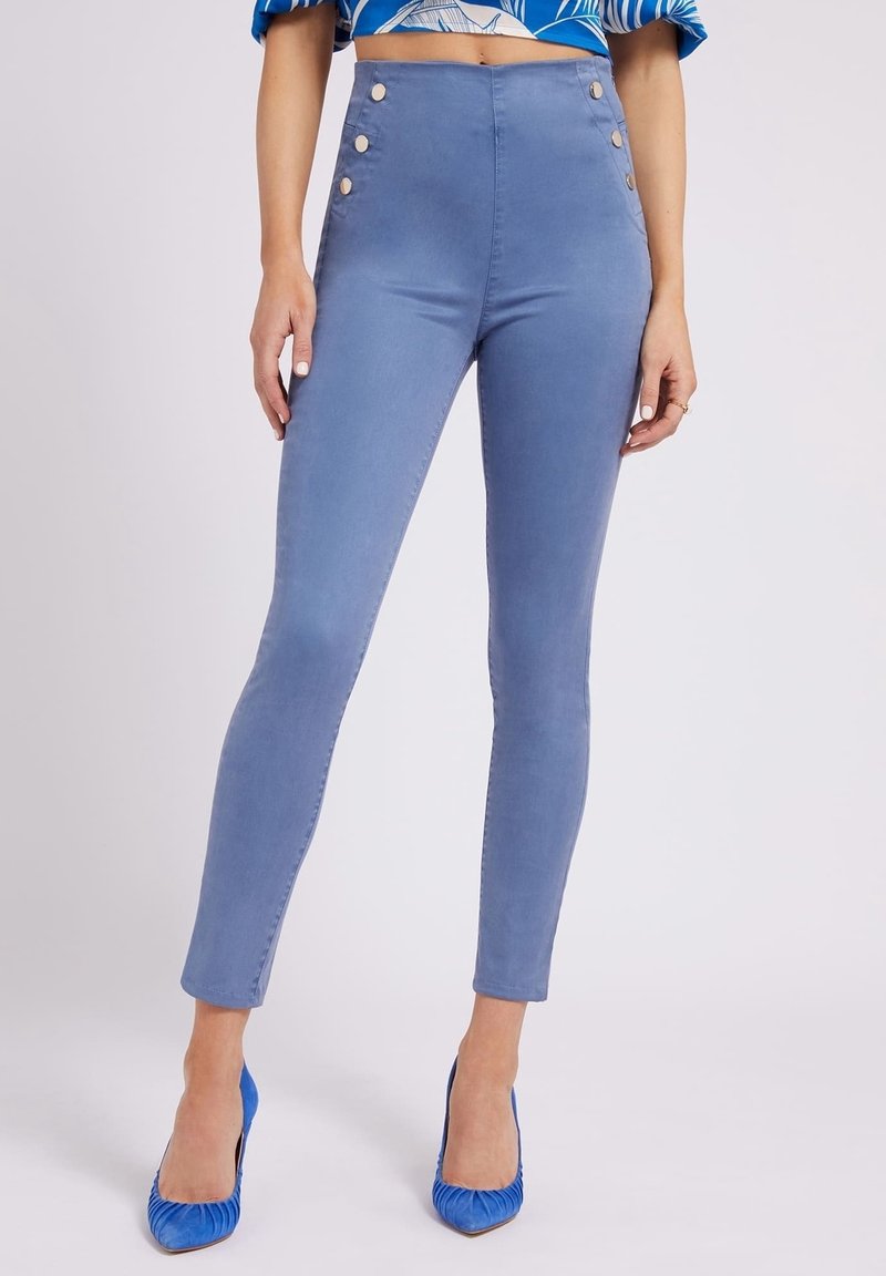 Vrouw in hooggetailleerde blauwe skinny broek met zijknopen, gecombineerd met blauwe hoge hakken en een cropped top met een patroon.