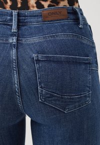 Jeans de mezclilla azul oscuro con un diseño ajustado, que cuentan con un bolsillo trasero visible y una etiqueta de marca de cuero marrón en la cinturilla.