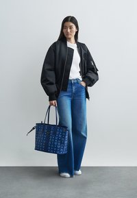 Juoda bombos tipo striukė, balti marškinėliai, mėlynos plačios kelnės ir mėlyna raštuota rankinė. Modelis stovi prieš neutralią foną.