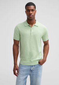 Jasnozielona koszulka polo z krótkim rękawem i żebrowanym dołem oraz kołnierzykiem, posiadająca trzy guziki i miękką teksturę. Połączona z jasnymi dżinsami.