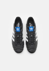 Sapatilhas de pele preta com riscas brancas e um logótipo azul da Adidas na língua. Parte superior texturizada e design de atacadores. Sola de borracha plana.