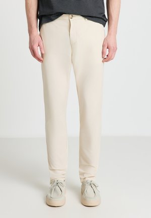 Homme portant un pantalon droit couleur crème et des chaussures à lacets beige clair sur un fond clair uni.
