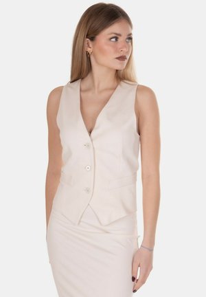 Giovane donna con capelli lunghi e lisci, indossa un gilet beige senza maniche e una gonna coordinata, guarda di lato su sfondo bianco.
