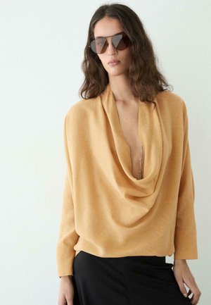 Žena nosící velký hořčicově žlutý přehozený svetr, černou sukni a velké čtvercové sluneční brýle na jednoduchém světlém pozadí.