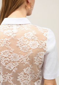 T-shirt polo blanc avec un dos en dentelle ornée de motifs floraux. Manches courtes et col classique. Texture en dentelle contrastant avec le tissu lisse.
