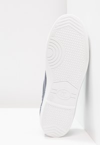 Suola in gomma bianca di una sneaker con superficie texturizzata, dotata di tomaia grigia scura e cuciture bianche lungo il lato. Dettagli delle dimensioni visibili.