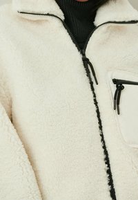 Giacca in pile crema con colletto alto, chiusura con zip e dettagli neri. Presenta una tasca con zip sul petto e un tessuto texturizzato.