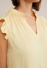 Femme portant un haut sans manches jaune clair avec des détails de volants aux épaules et un petit col volanté, dévoilant la partie supérieure de la poitrine et le cou.