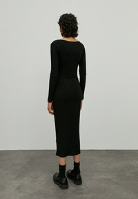 Schwarzes, geripptes Langarmkleid mit taillierter Silhouette, das bis zur Mitte der Wade reicht. Kombiniert mit klobigen schwarzen Schuhen und Socken. Glatte Textur.