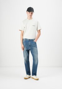 Hvid kortærmet t-shirt med sort logo, blå jeans og hvide sneakers med tan sål. Kasket i sort kompletterer det afslappede outfit.