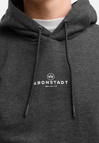 Felpa grigia scura con cappuccio, caratterizzata da un logo bianco centrale "KRONSTADT" e cordini con punte nere. Tessuto morbido.