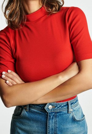 Vrouw met gekruiste armen, gekleed in een rood geribbeld topje met korte mouwen en blauwe denim high-waisted jeans, ingekort bij de hals en taille.