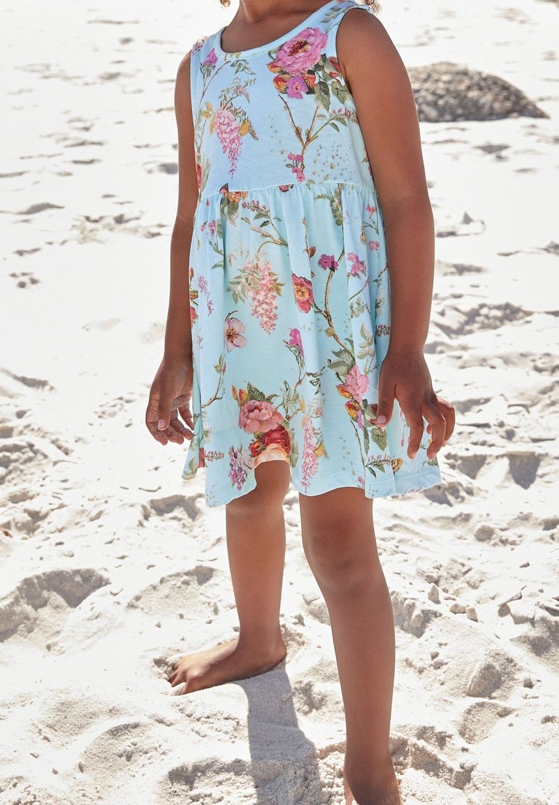 Robe sans manches bleu clair à motif floral rose et vert, jupe fluide et taille froncée, portée pieds nus sur un terrain sablonneux.
