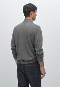Grå stickad tröja med långa ärmar, ribbade muddar och nederkant, bärs över mörka denimjeans. Bakifrån visar den en minimalistisch design.