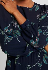 Blommig klänning i marinblått tyg med puffärmar, med gröna och rosa botaniska tryck. Detaljer inkluderar knäppta manschetter och en avslappnad passform.