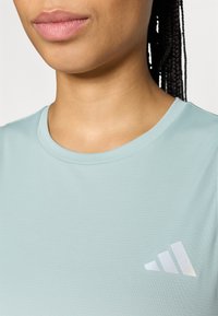 Hele sinine sportlik särk ümarate kaelusega, tekstureeritud kangas ja peegeldav Adidas logo, mis koosneb kolmest kaldus triibust.