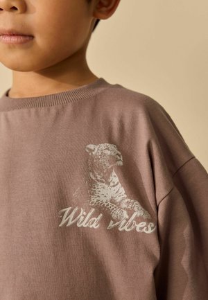 Kind trägt einen mauvefarbenen Pullover mit einer Zeichnung eines ruhenden Leoparden und dem Schriftzug "Wild vibes" auf der Brust.