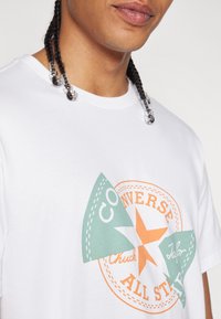 Camiseta de algodón blanca que presenta un logo circular naranja y verde de Converse All Star con una estrella y detalles de costura en el pecho.