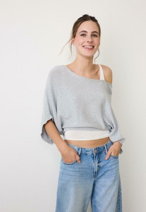 Donna sorridente con capelli castani, maglione grigio largo con spalle scoperte, canottiera bianca e jeans azzurri a vita alta con le mani nelle tasche.