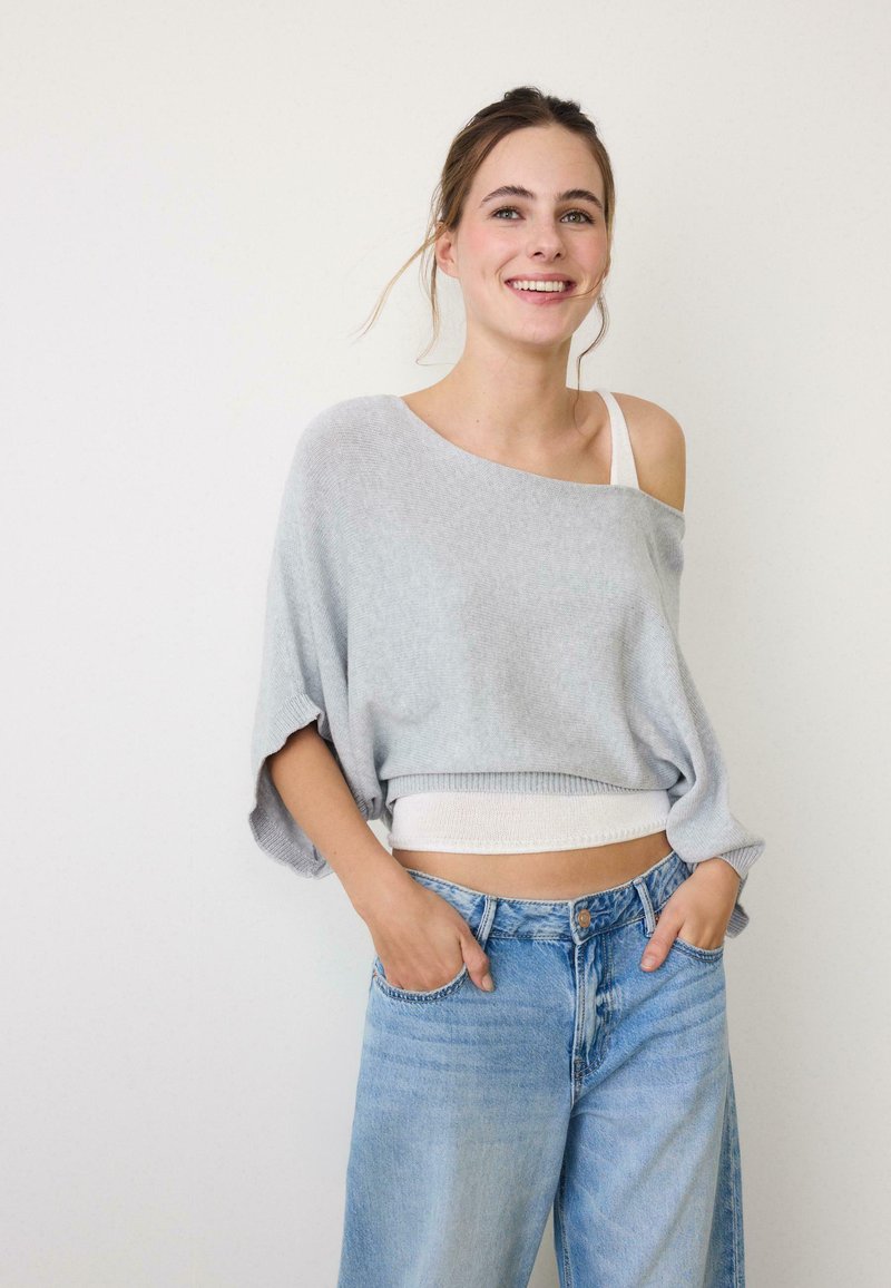 Smilende kvinde med brunt hår iført en løs, grå off-shoulder sweater, hvid tanktop og lyseblå jeans med høj talje og hænderne i lommerne.