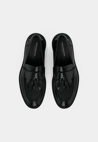 Mocassins en cuir noir avec ornement à pompon, texture lisse, bout arrondi et couture détaillée le long des bords. Nom de la marque visible à l'intérieur.