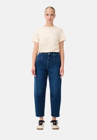 T-shirt di cotone beige chiaro con maniche corte, abbinato a jeans high-waisted in denim blu scuro con tasche frontali. Scarpe stringate nere e crema.