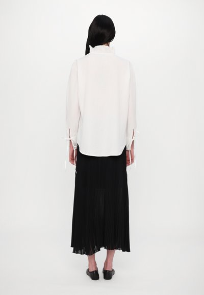 Weiße Bluse mit Rüschensaum und Bindebändern an den Ärmeln, kombiniert mit einem langen schwarzen Faltenrock und schwarzen Slippern.