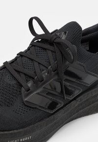 Černá pletená atletická bota s texturovanou podrážkou, černé tkaničky uvázané do mašle a lesklý černý design se třemi pruhy na boku.
