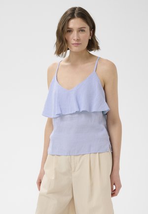 Femme aux cheveux courts bruns portant un haut sans manches bleu clair à volants et un pantalon beige taille haute à jambes larges, se tenant devant un fond gris uni.