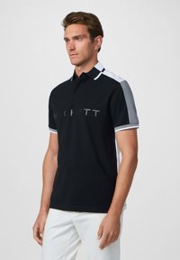 Camisa polo negra con un cuello y mangas a rayas en blanco y gris. Presenta un destacado logo "HACKETT" en un tono más claro en la parte delantera.