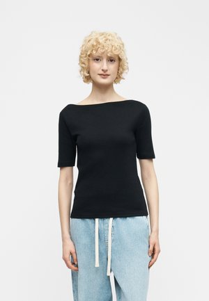 Mujer rubia con cabello rizado, vestida con una camiseta negra de manga corta y cuello barco, y pantalones vaqueros azul claro con cordones blancos, de pie contra un fondo blanco.