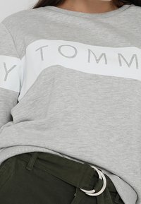 Szara bluza z białym poziomym paskiem z napisem "TOMMY" w kontrastującym kolorze. Posiada okrągły dekolt i ściągacz na dole.