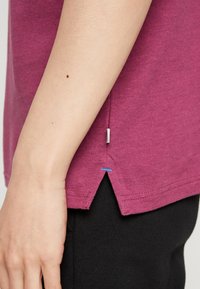 Vinröd bomulls-T-shirt med en sidofåra, kontrasterande blå sömmar och en strukturerad tygkvalitet. Bärs över svarta byxor.