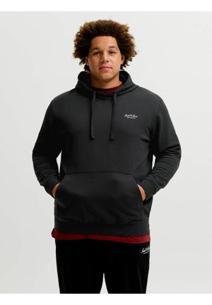 Homme aux cheveux bouclés portant un sweat à capuche noir et un pantalon noir, les mains dans la poche du sweat à capuche, sur un fond clair uni.