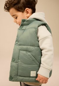 Zielona kamizelka z wypełnieniem, o pikowanym wzorze, z zatrzaskami i przednią kieszenią. Nosi ją się na szarym hoodie z kontrastowymi mankietami.