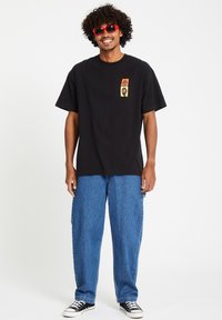Black cotton t-shirt with a front pocket graphic. Paired with loose blue denim pants and black sneakers. Red sunglasses complete the look.