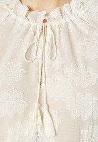 Blouse crème avec motif floral texturé, encolure nouée avec deux pompons tressés et un bord de col subtilement froncé.