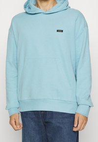 Felpa leggera blu chiaro in materiale morbido, con tasca a marsupio, polsini a costine e un piccolo patch logo nero sul petto.