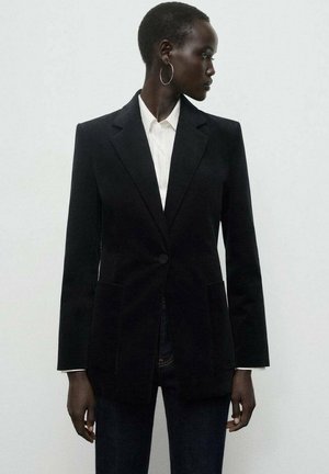 Blazer negro a medida con solapa muesca, cierre de un solo botón y dos bolsillos frontales; combinado con una camisa blanca de cuello por debajo.