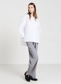 Woman wearing a loose white button-up shirt, gray pinstripe trousers, and black loafers, standing with hands in pockets against a plain background.