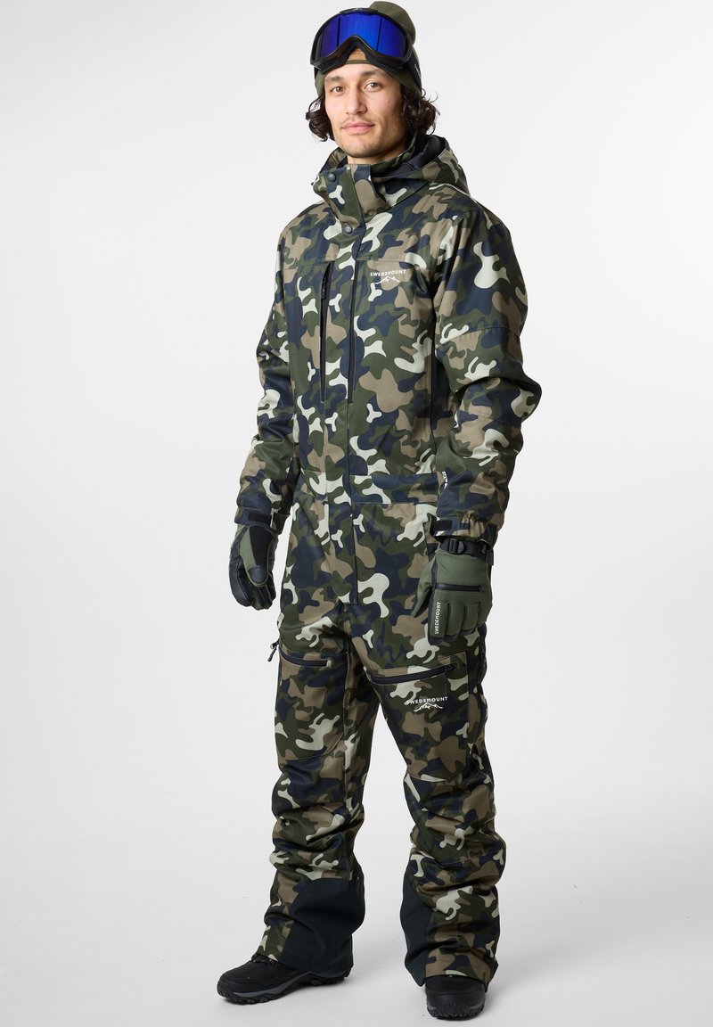 Vihreä, beige ja musta camo-talvitakki. Sisältää hupun, vetoketjut edessä ja hanskat. Päällä suojalasit ja lumitossut. Teksturoitua kangasta.