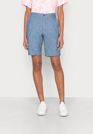 Person trägt mittellange blaue Chambray-Shorts, ein pinkes Batik-Shirt und weiße Sneaker und steht vor einem schlichten Hintergrund.