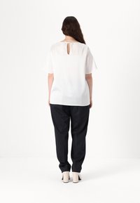 Blusa bianca trasparente a maniche corte, con dettaglio a chiave sul retro, abbinata a pantaloni neri a righe e scarpe con tacco spesso color beige.