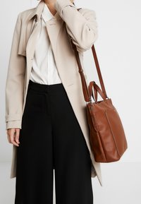 Manteau beige sur une chemise blanche, pantalon noir à larges jambes, tenant un sac à main en cuir marron avec une surface texturée et des coutures contrastées.