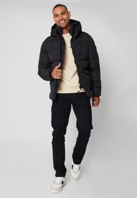 Kurtka puchowa, czarna, z kapturem i zamek błyskawiczny; noszona na beżowej bluzie i zestawiona z czarnymi spodniami cargo oraz białymi trampkami.