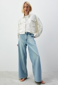 Hvid, cropped, button-up denimjakke med sølvknapper og to frontlommer, parret med oversized lyseblå jeans og sandaler.