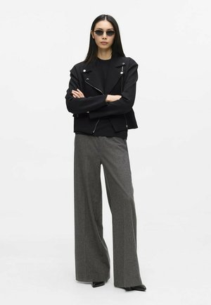 Frau mit glattem schwarzen Haar, die kleine schwarze Sonnenbrillen, eine schwarze Jacke, ein schwarzes Oberteil, weite graue Hosen und schwarze Schuhe trägt und mit verschränkten Armen steht.